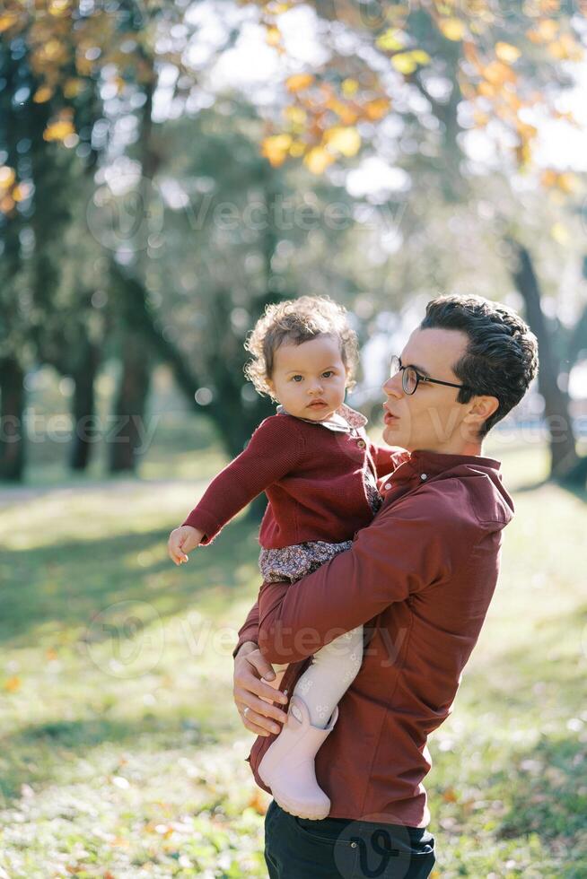 Dad is talking to a little girl in his arms in a sunny park photo