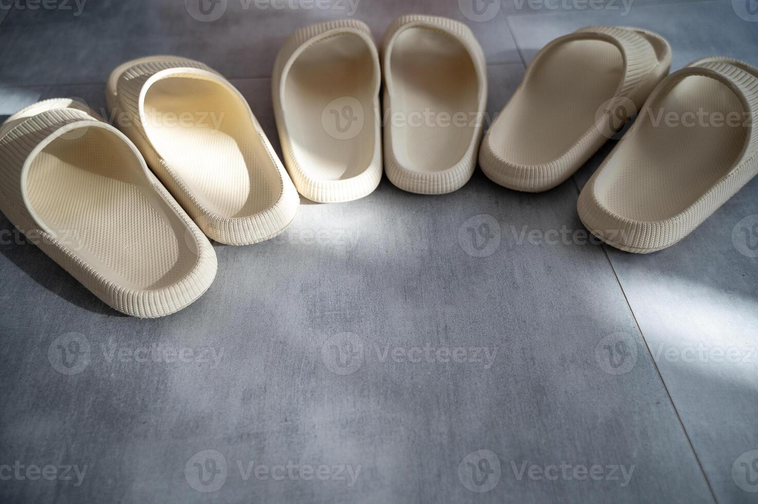 Three pairs of beige rubber slippers stand on a gray granite floor. A ray of sunlight falls on them. The slippers stand in a row, in a semicircle. They are the same size, viewed from above. Close-up photo