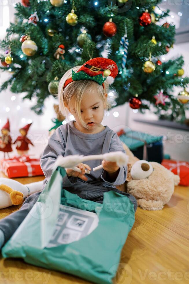 Little girl in a hoop with a gnome hat examines a hoop with cat ears, sitting by the Christmas tree photo