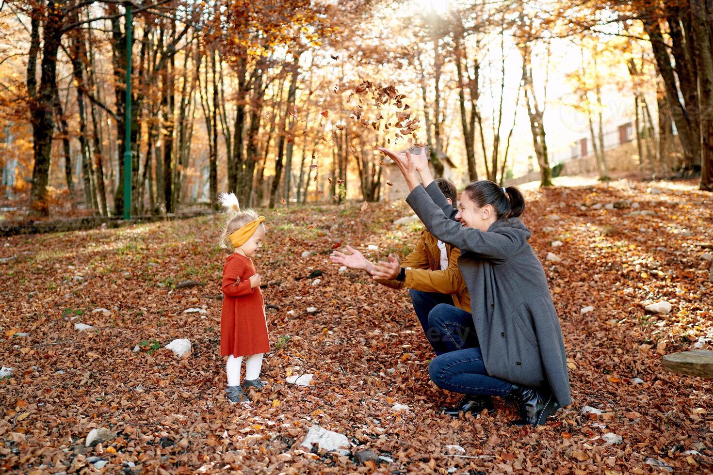 Little girl stands in front of mom and dad squatting and throwing up leaves in the autumn park photo
