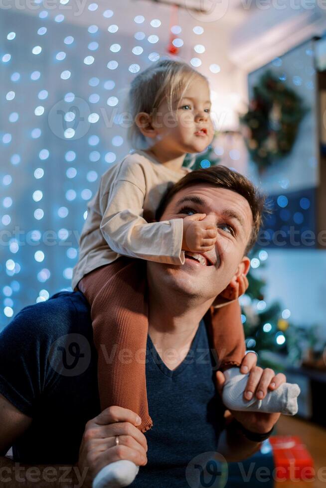 Little girl touching dad nose while sitting on his shoulders near the decorated Christmas tree photo