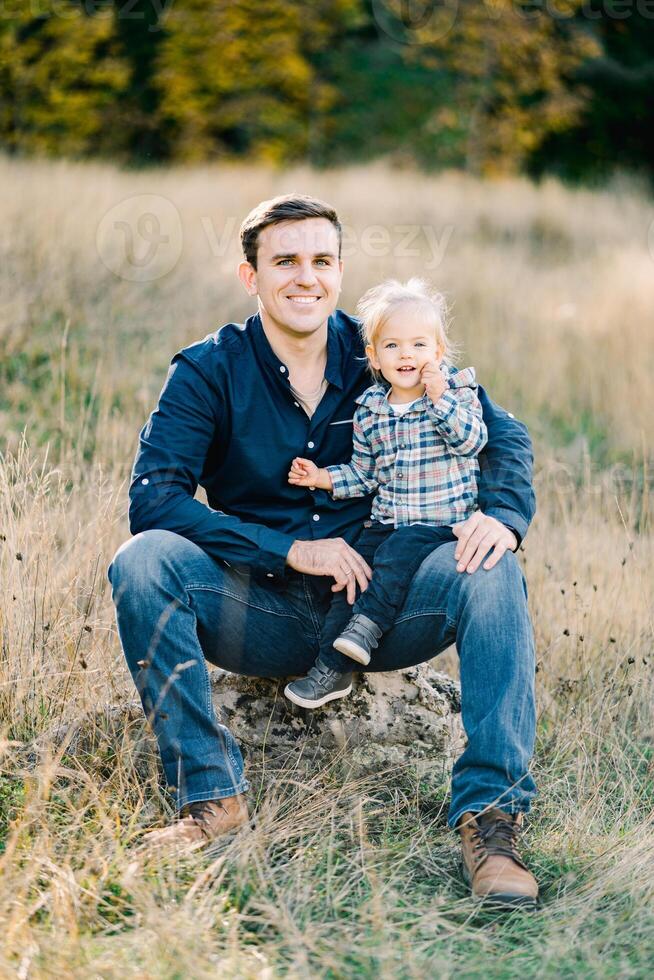 Little girl sits on her father lap on the lawn at the edge of the forest photo