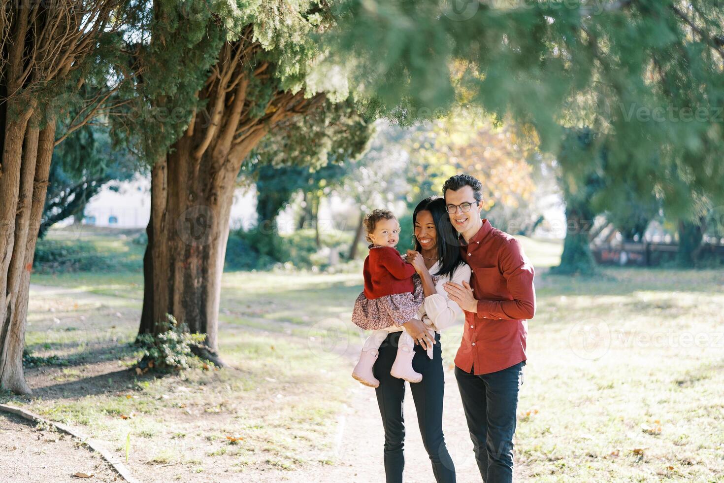 Smiling dad hugging mom from behind with a little girl in her arms near a tree photo