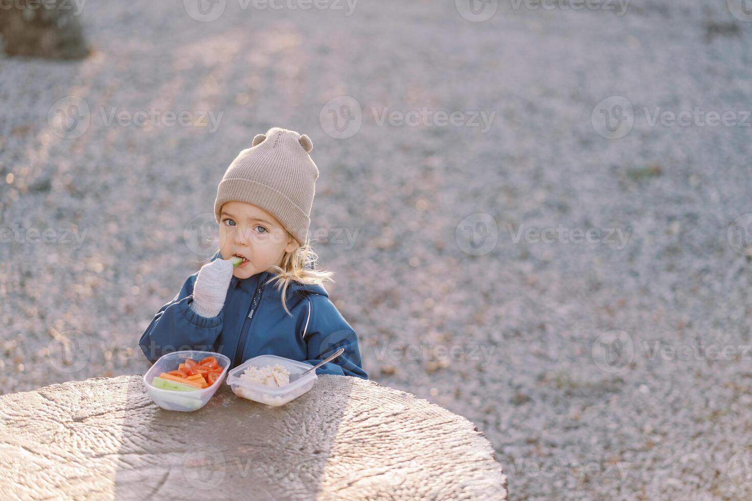 Little girl eats a piece of cucumber while standing behind a large tree stump with a lunchbox photo