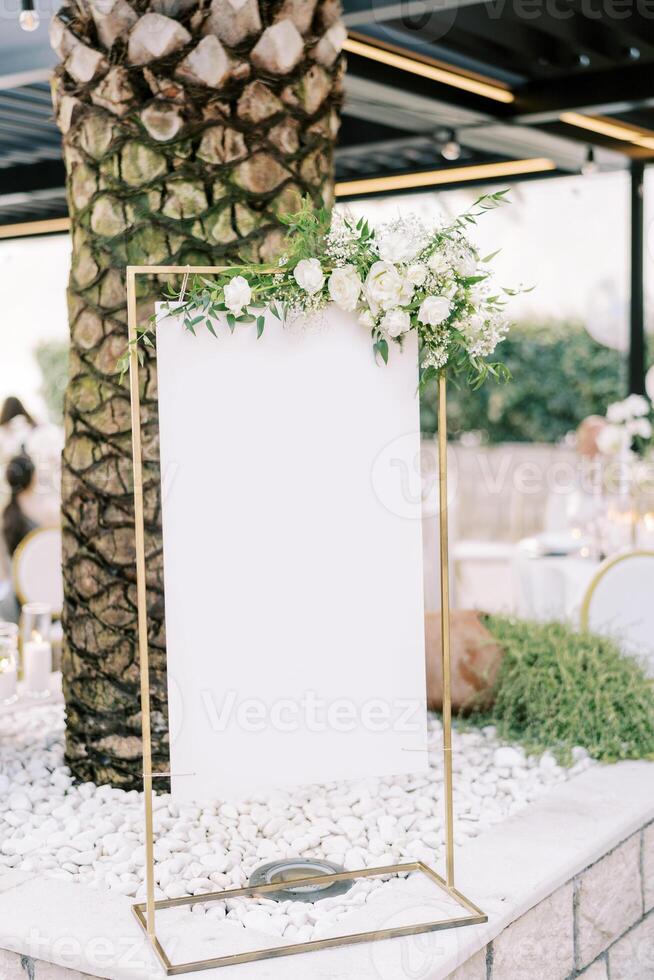 Empty sign on a stand decorated with white flowers stands near a palm tree photo