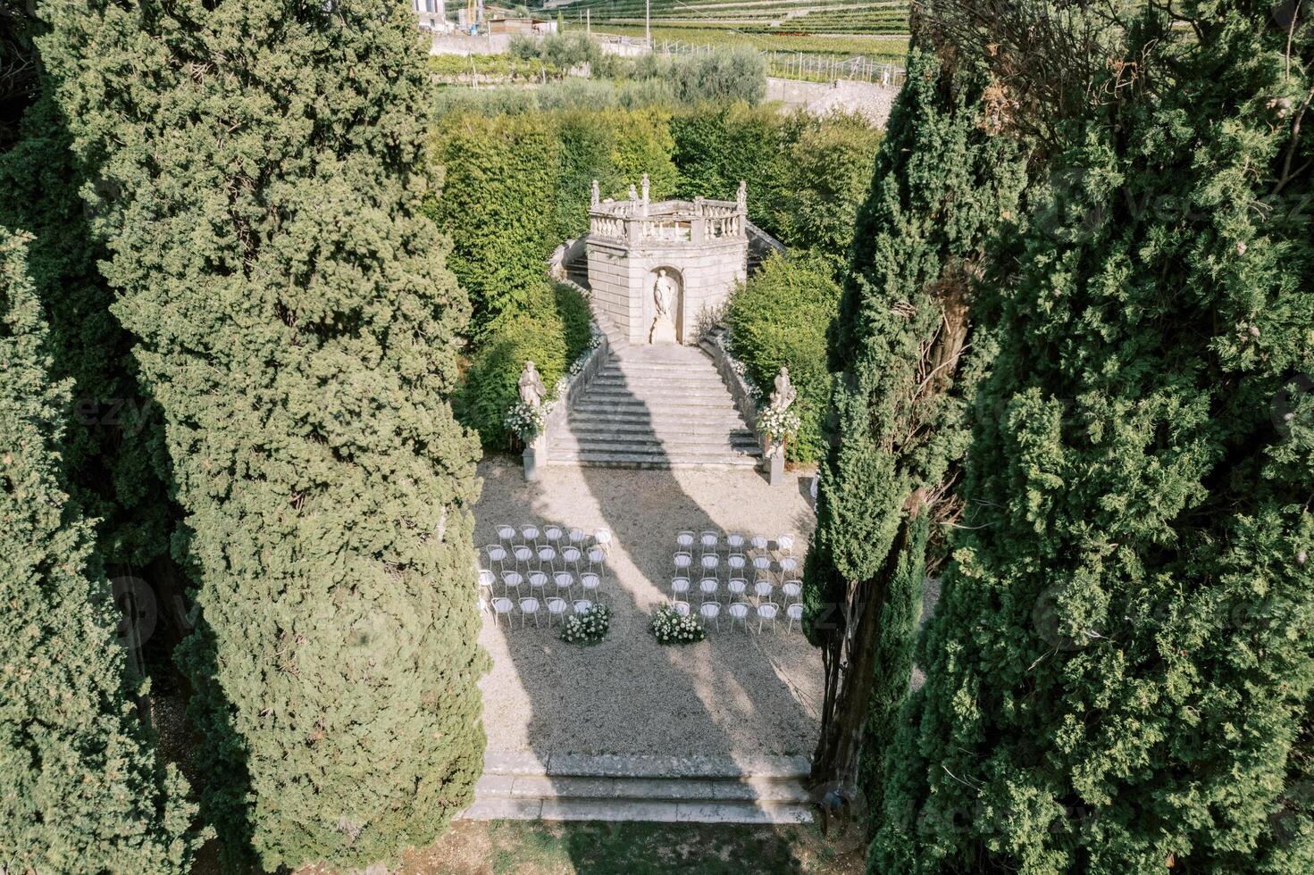 Chairs stand in rows on a park alley near steps decorated with flowers in front of a statue of a woman. Top view photo