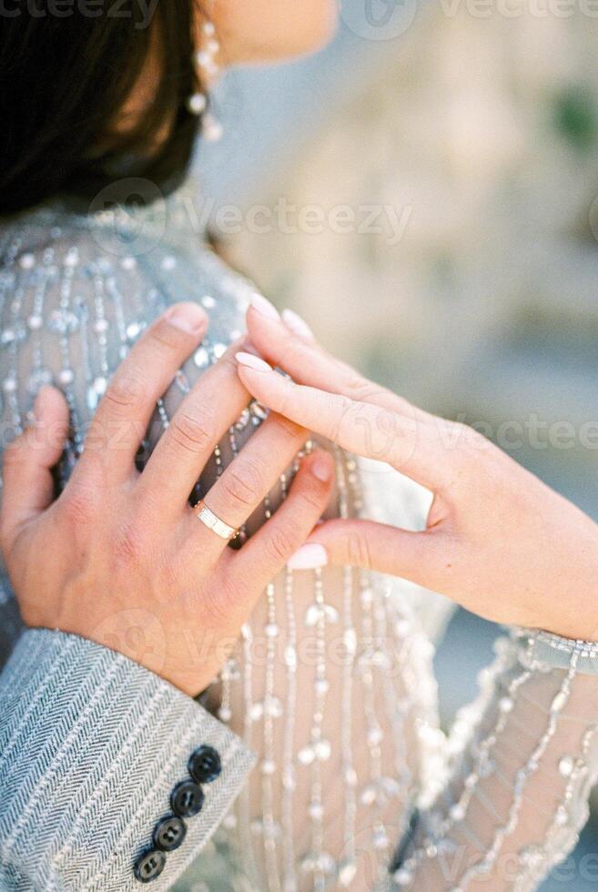 Bride put her hand on the hand of groom hugging her shoulder. Back view. Cropped photo