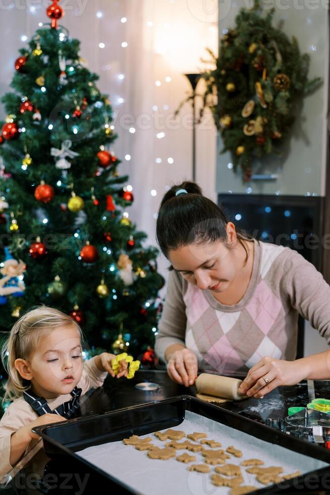 Little daughter looks at the cookies on the tray while mom rolls out the dough with a rolling pin photo