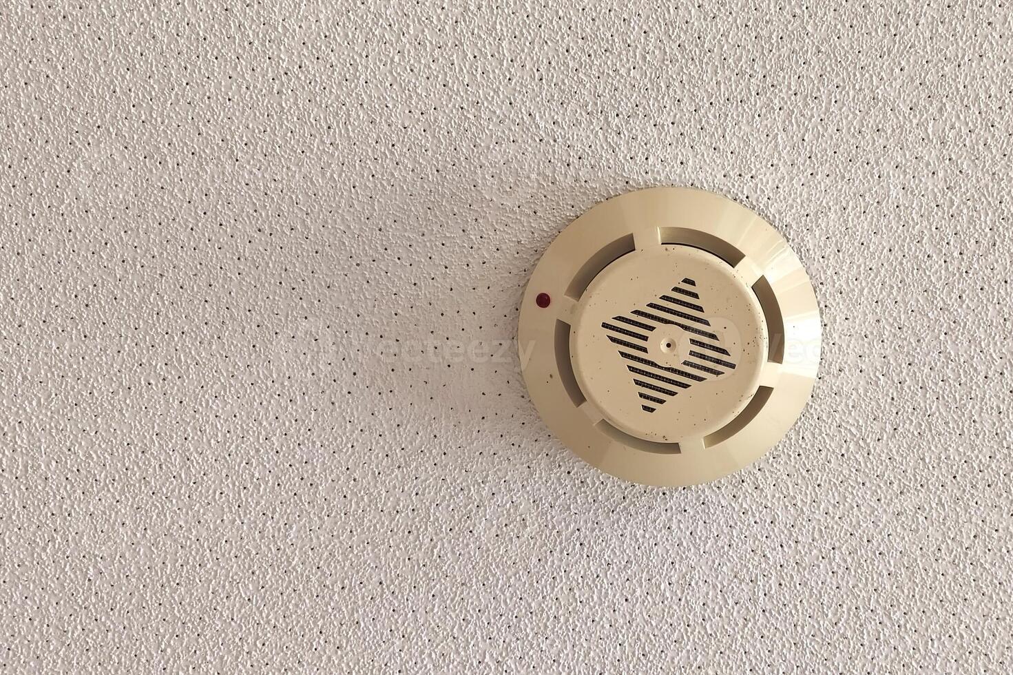 A smoke detector is mounted on the ceiling of a living space, functioning as a crucial safety device to detect fire hazards. It features a circular design with a grill and indicator light. photo