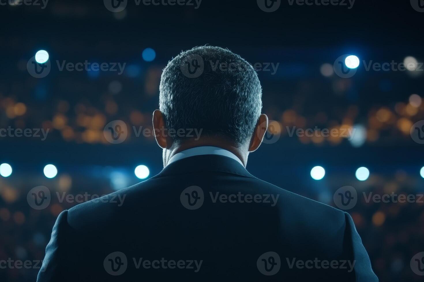 Speaker addresses large audience at nighttime event in an arena filled with bright lights and eager attendees photo