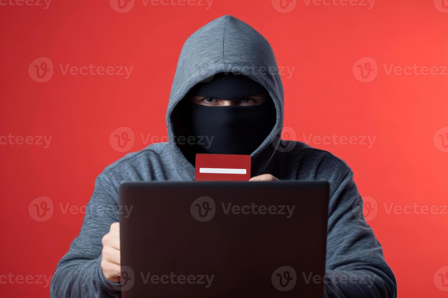 Cybercriminal wearing a hoodie engages in identity theft while using a laptop against a red background photo