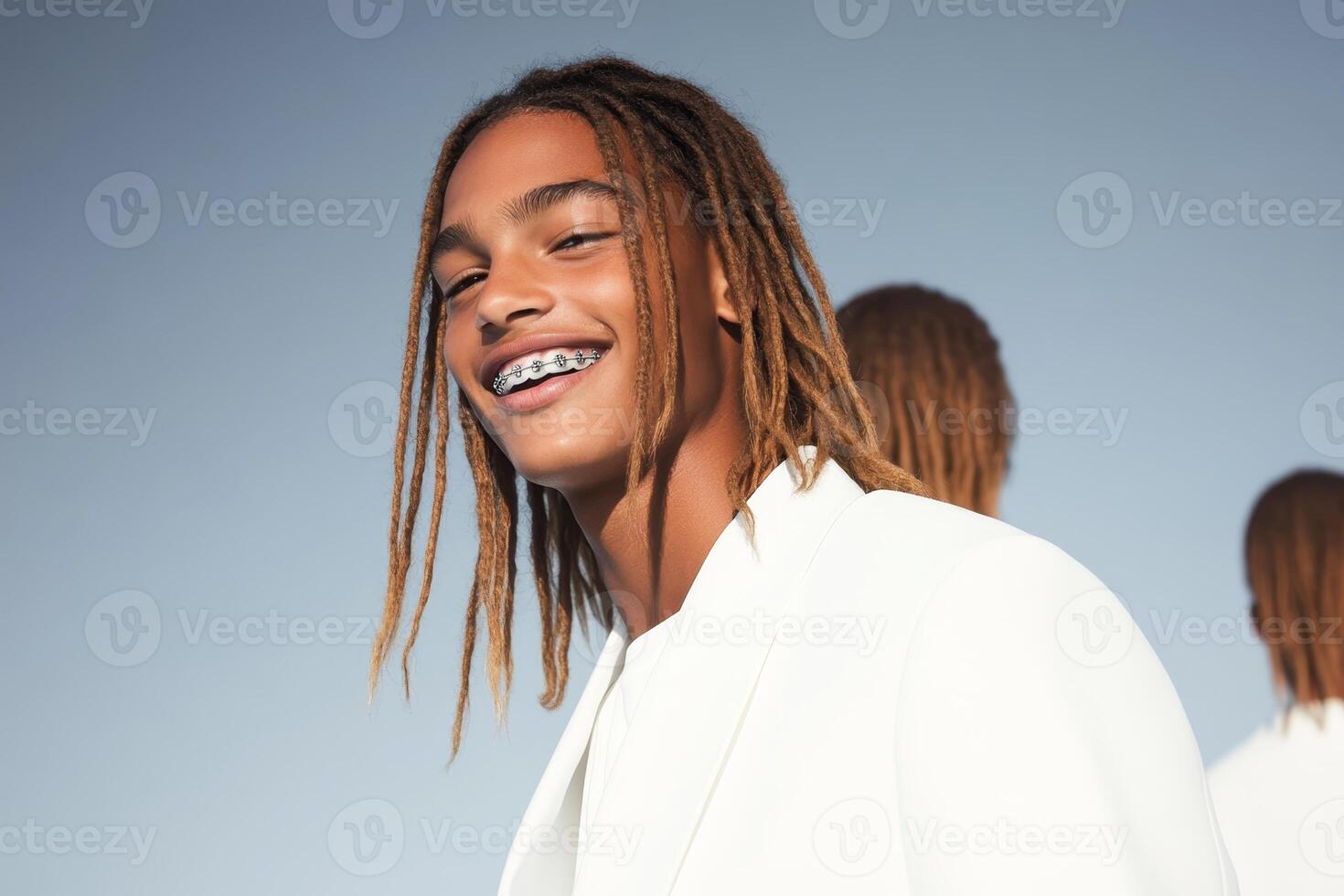 Young man with dreadlocks wearing a stylish white outfit smiles confidently against a clear sky backdrop in a modern setting photo