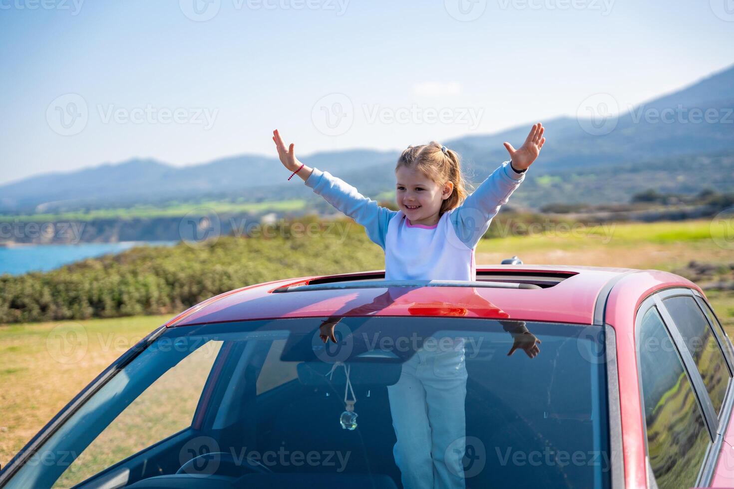 Little girl joyfully sticks out of car sunroof with raised hands against the backdrop of mountains in Northern Cyprus. The image symbolizes freedom, adventure, and the excitement of travel with child. photo