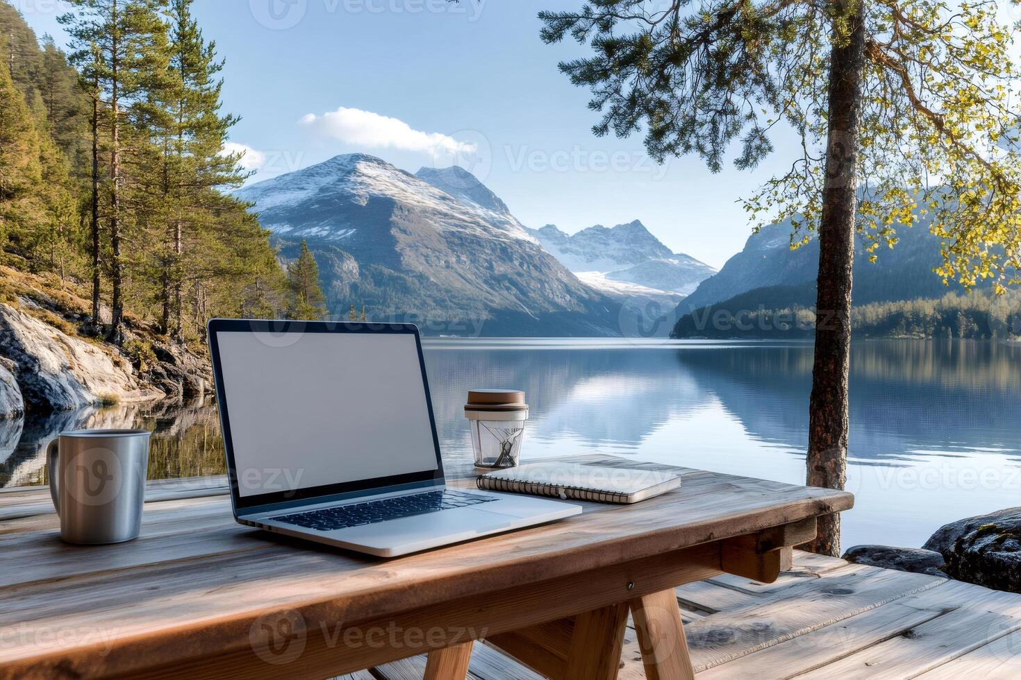 Laptop with blank screen, coffee and notebook placed on wooden table enjoying mountain lake view photo
