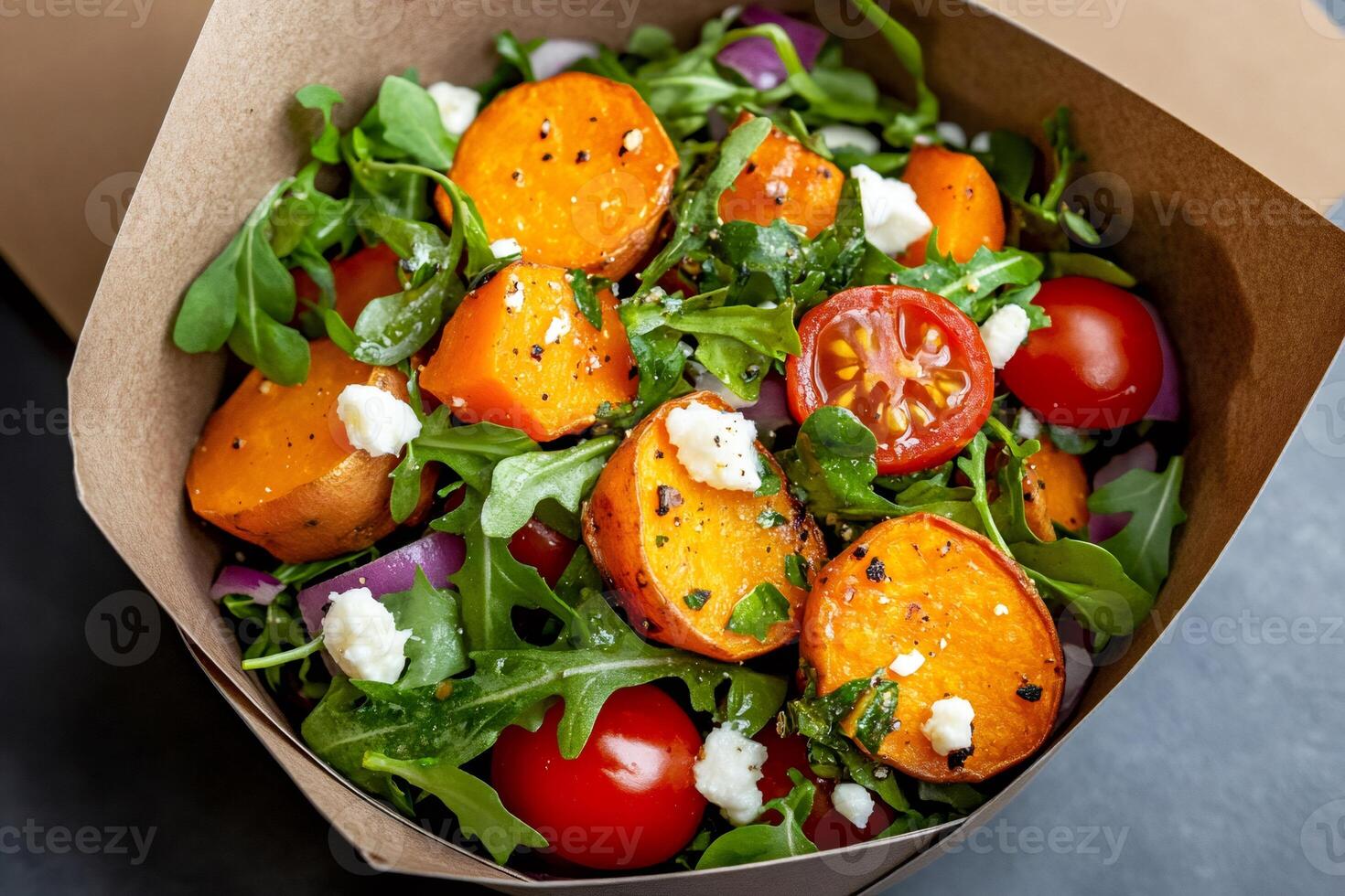 Healthy salad with baked sweet potatoes, arugula, tomatoes and feta cheese in a cardboard bowl photo