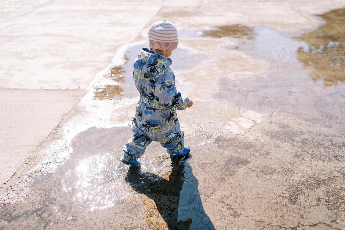 Little girl in rubber boots walks through a puddle on the pavement. Back view photo