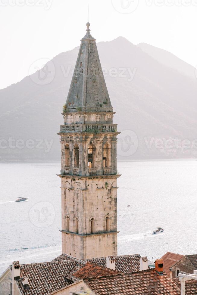 High spire of the ancient bell tower of the Church of St. Nicholas against the backdrop of boats sailing on the sea. Perast, Montenegro photo