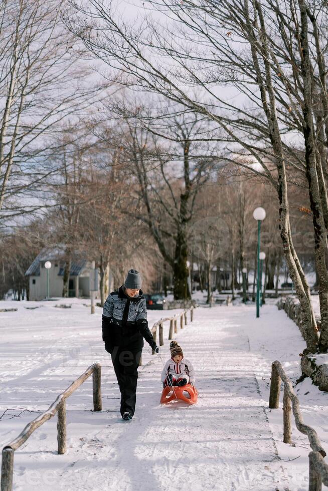 Smiling mother carries a small child on a sled on a path in the park photo