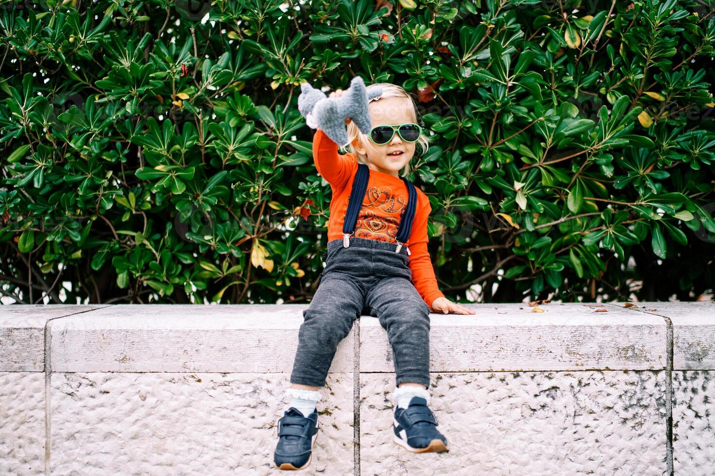 Little girl in sunglasses with a soft toy in her hand sits on a stone fence near a green bush photo