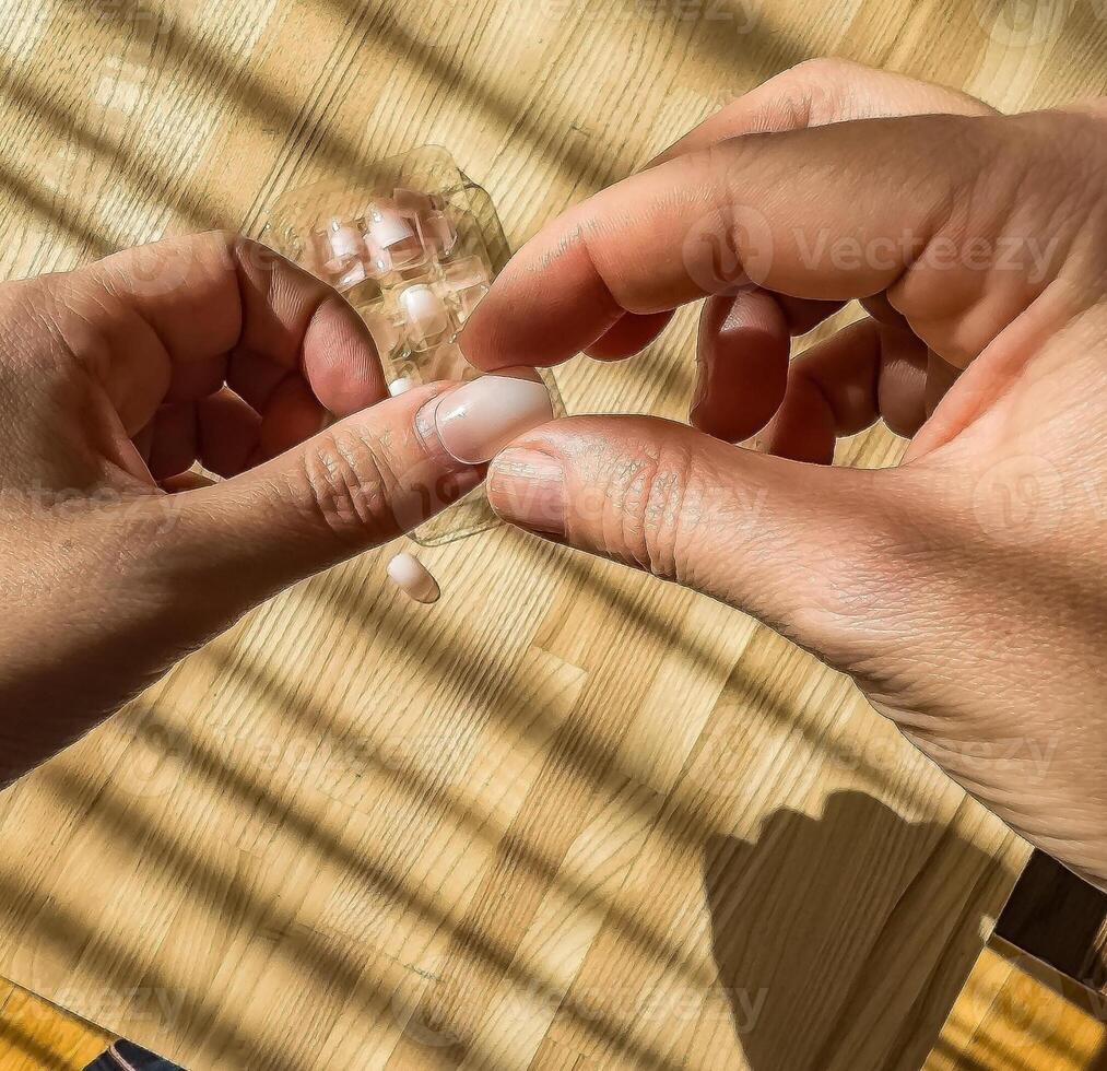 Close-up of hands applying nude-colored press-on nails on a wooden surface. High quality photo