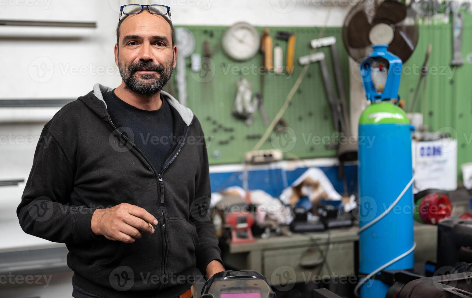 Portrait of a blacksmith during a break in a welding session. He takes the opportunity to smoke a cigarette, the welder's helmet can be seen in the bottom right. Blurred background with workshop. photo