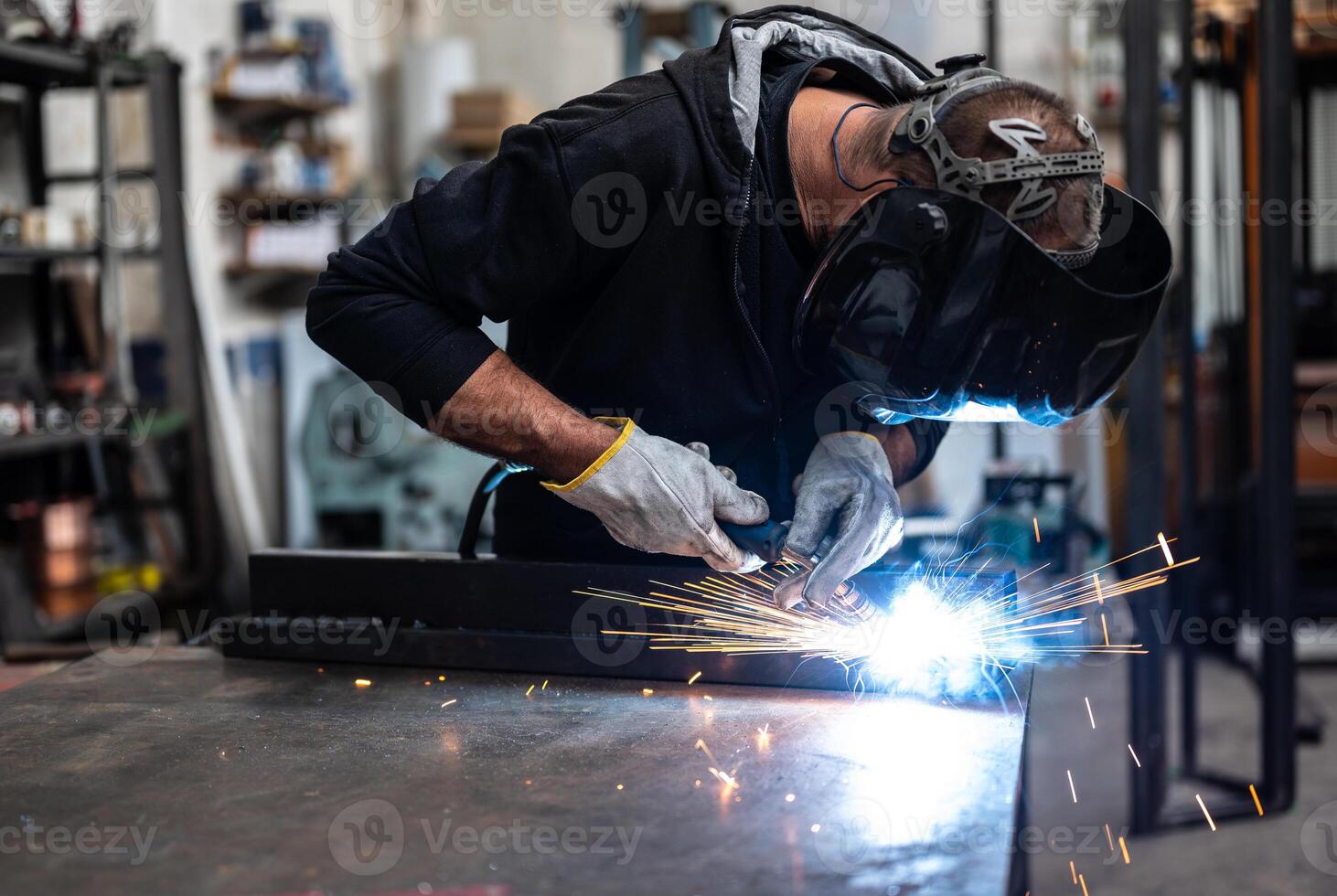 Performing a weld , the man in the welding mask is positioned with the welding machine on the welding point. When he activates it, a blue glow with smoke and orange sparks illuminates the scene photo