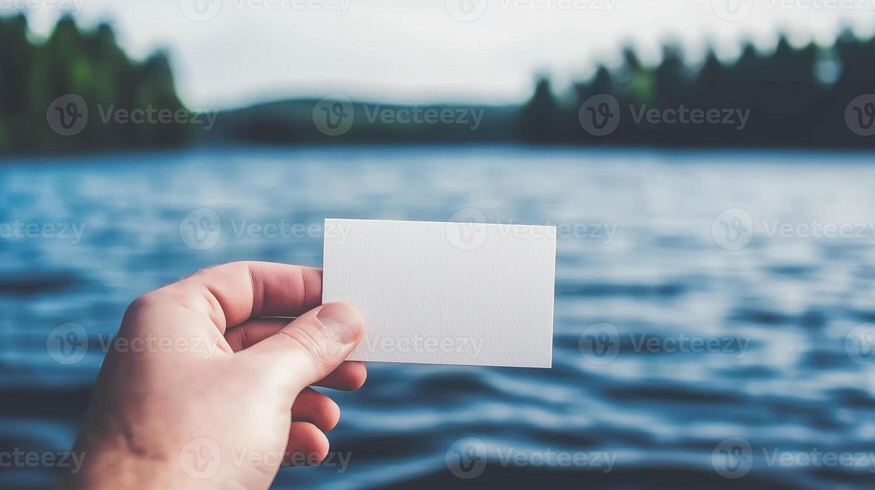 Person Holding Blank Business Card Near Calm Water with Green Hills in Background Under Bright Blue Sky photo