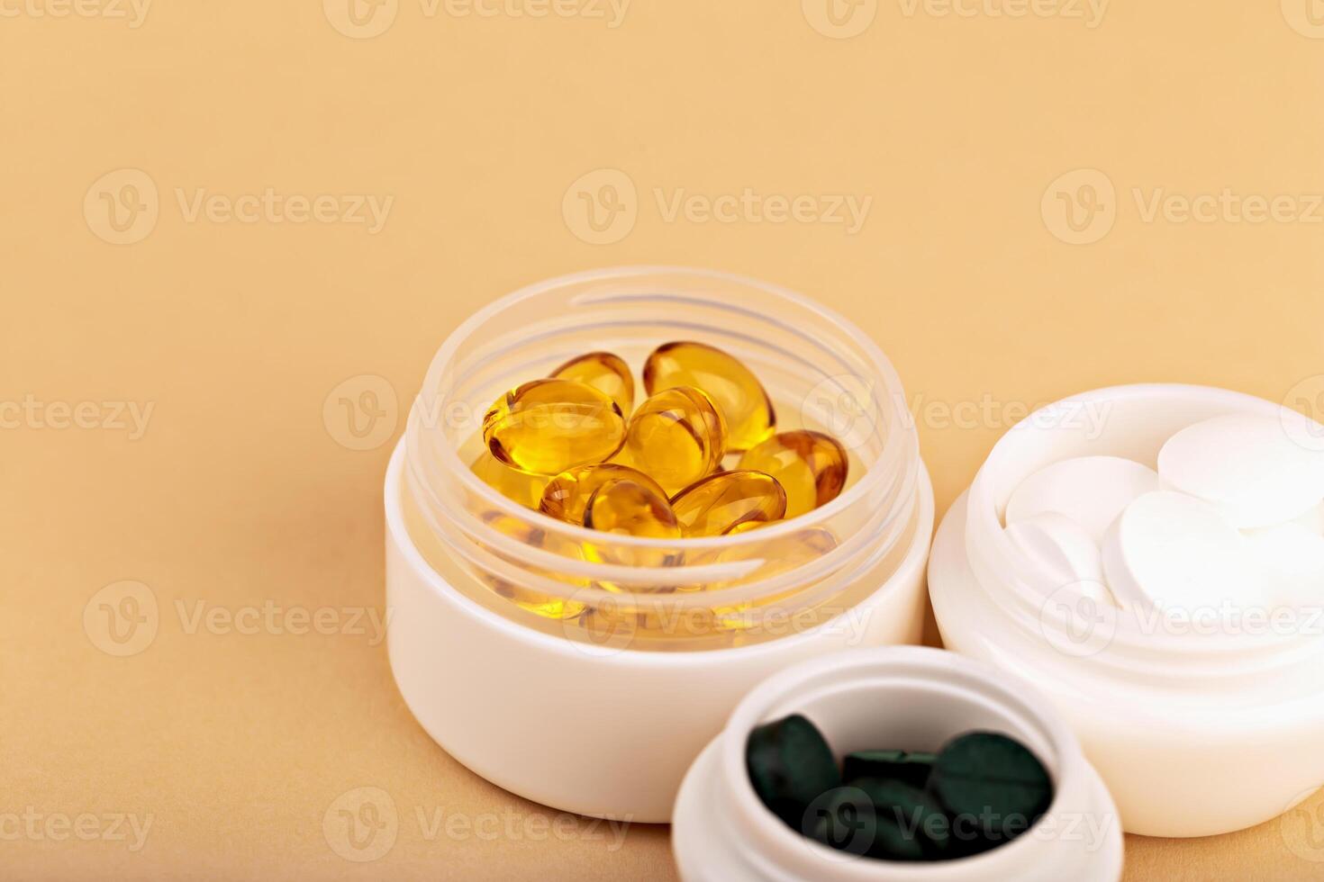 Three open white jars with yellow softgel capsules, white tablets and green spirulina pills on beige background. Concept of healthcare, beauty or supplement product. Open supplement jars with capsules photo