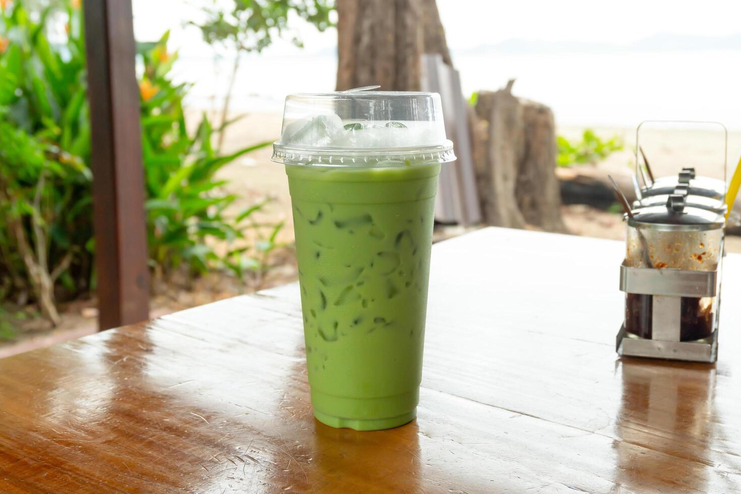 Cold drink of iced matcha latte green tea with condensed milk in transparent plastic drinking glass on table of out door cafe near the sea photo