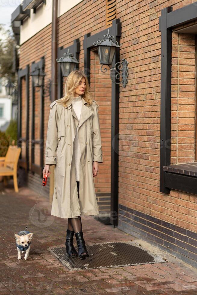 Woman walking her small dog on a city street in a stylish trench coat during a sunny day photo
