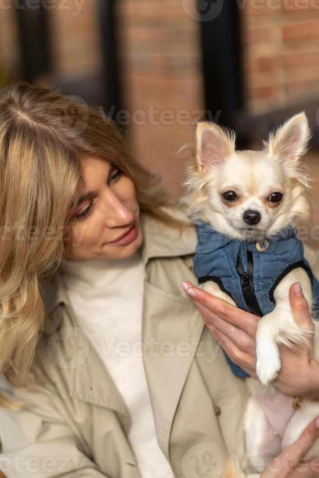 Woman holds small dog in denim jacket while smiling warmly at the camera in a cozy outdoor setting photo