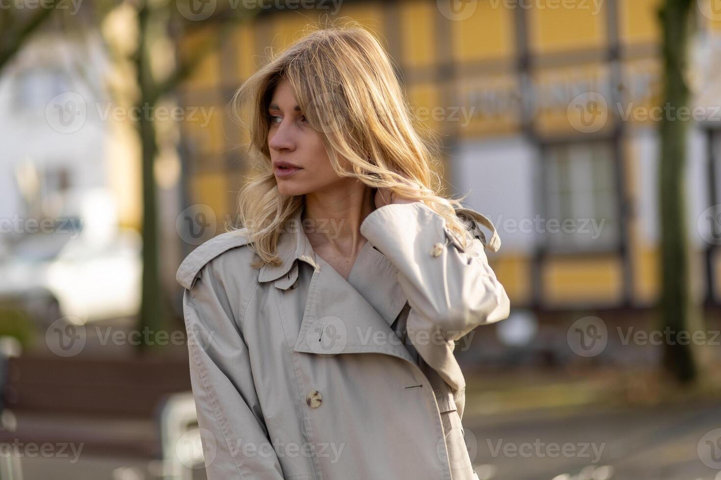 Woman in a light trench coat poses outdoors in a quaint town setting during the golden hour photo