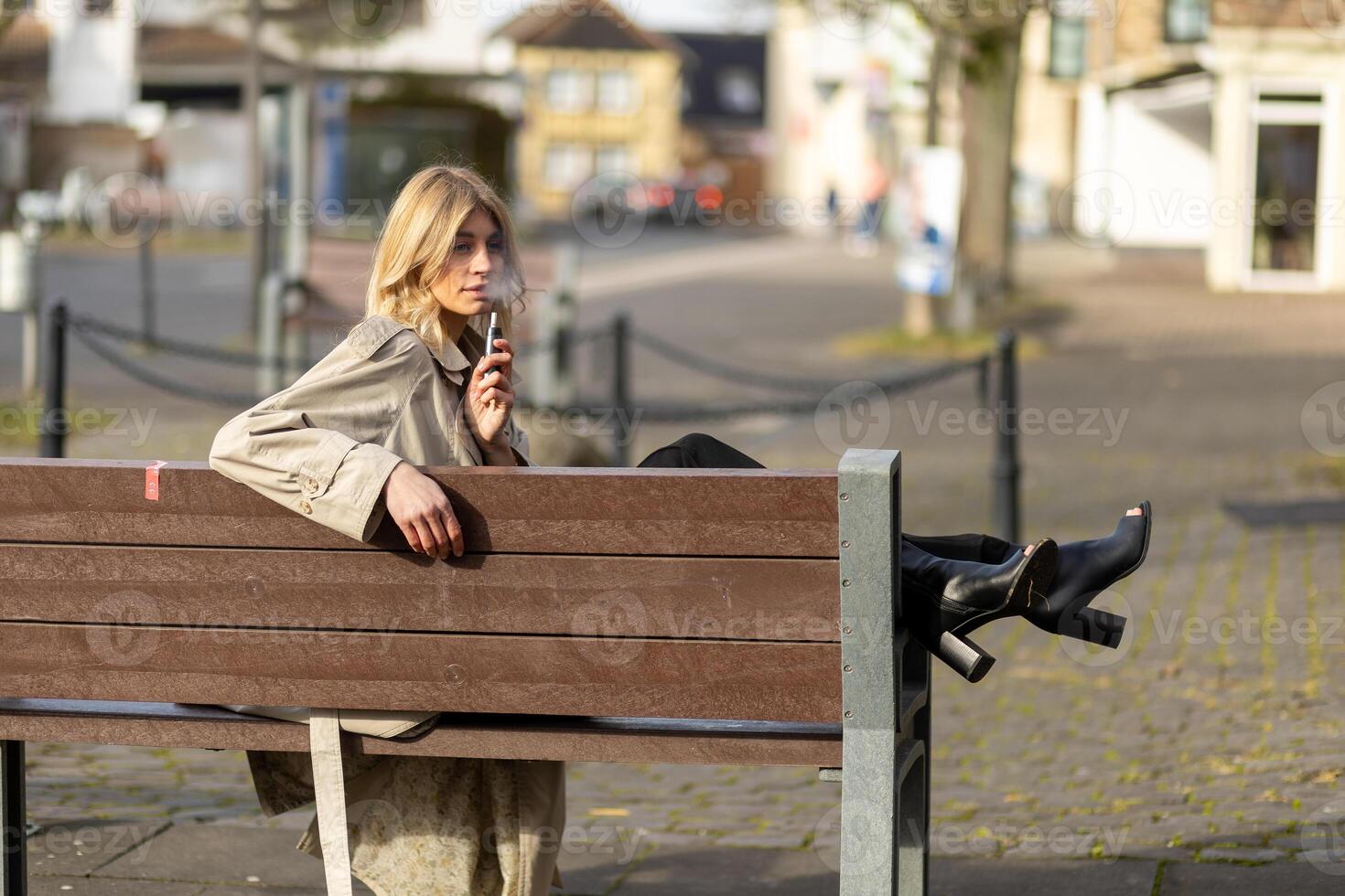 Young woman sitting on a park bench casually using a vape device in an urban environment during daylight photo