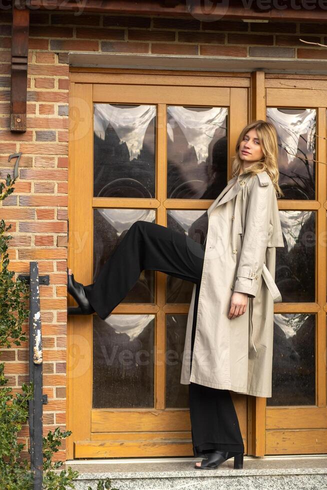 Woman posing stylishly in a trench coat with one leg on a doorstep in an urban setting during the day photo