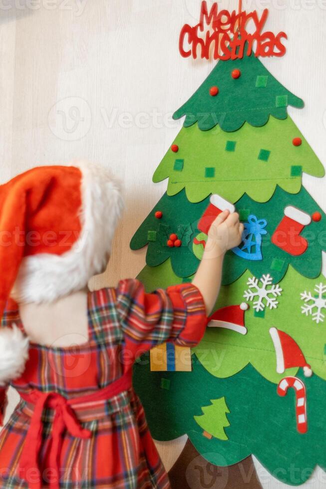 A young child decorates a colorful felt Christmas tree with ornaments while wearing a festive red hat in a cozy indoor setting photo