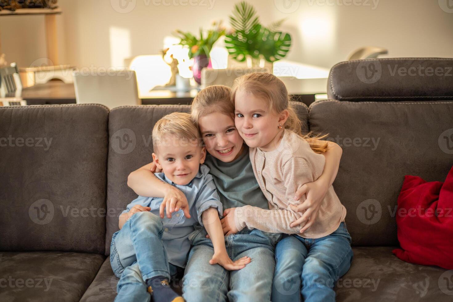 portrait of three children at home. brother and sisters sit on the couch hugging and smiling photo