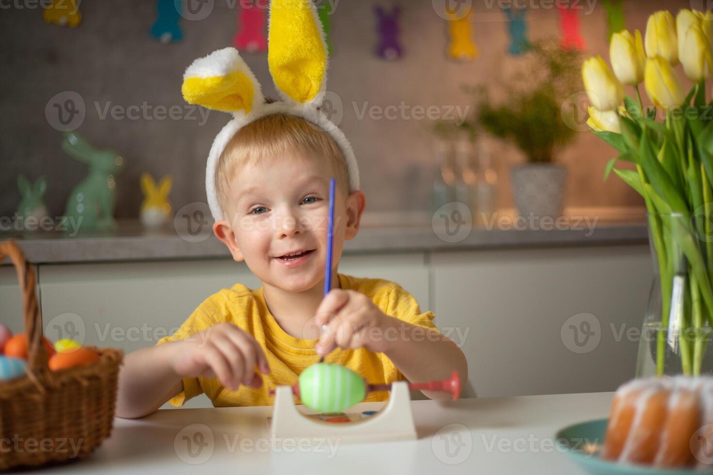 A little boy wearing bunny ears prepares painted Easter eggs for Easter while sitting at the kitchen table. Happy easter photo