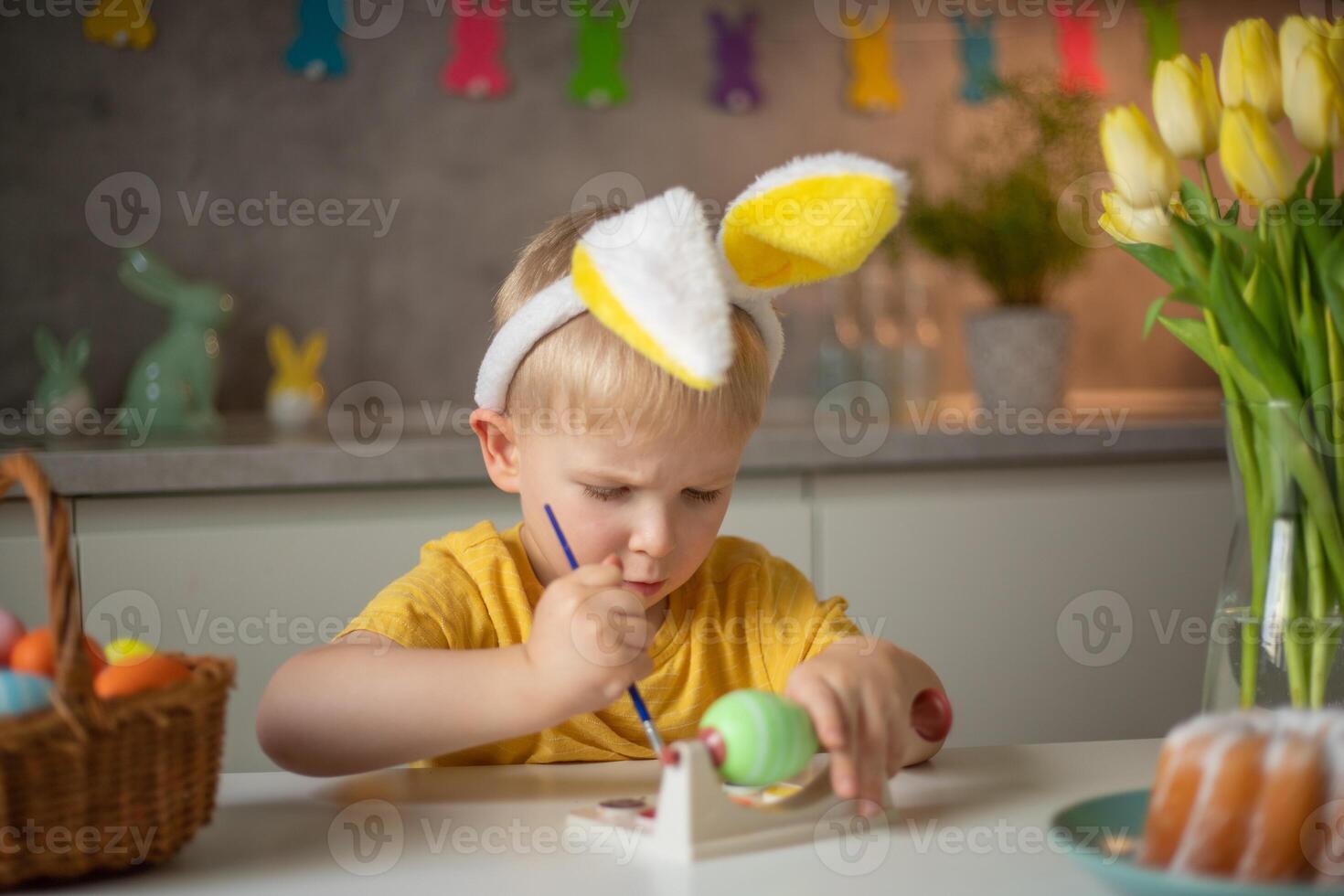 A little boy wearing bunny ears prepares painted Easter eggs for Easter while sitting at the kitchen table. Happy easter photo