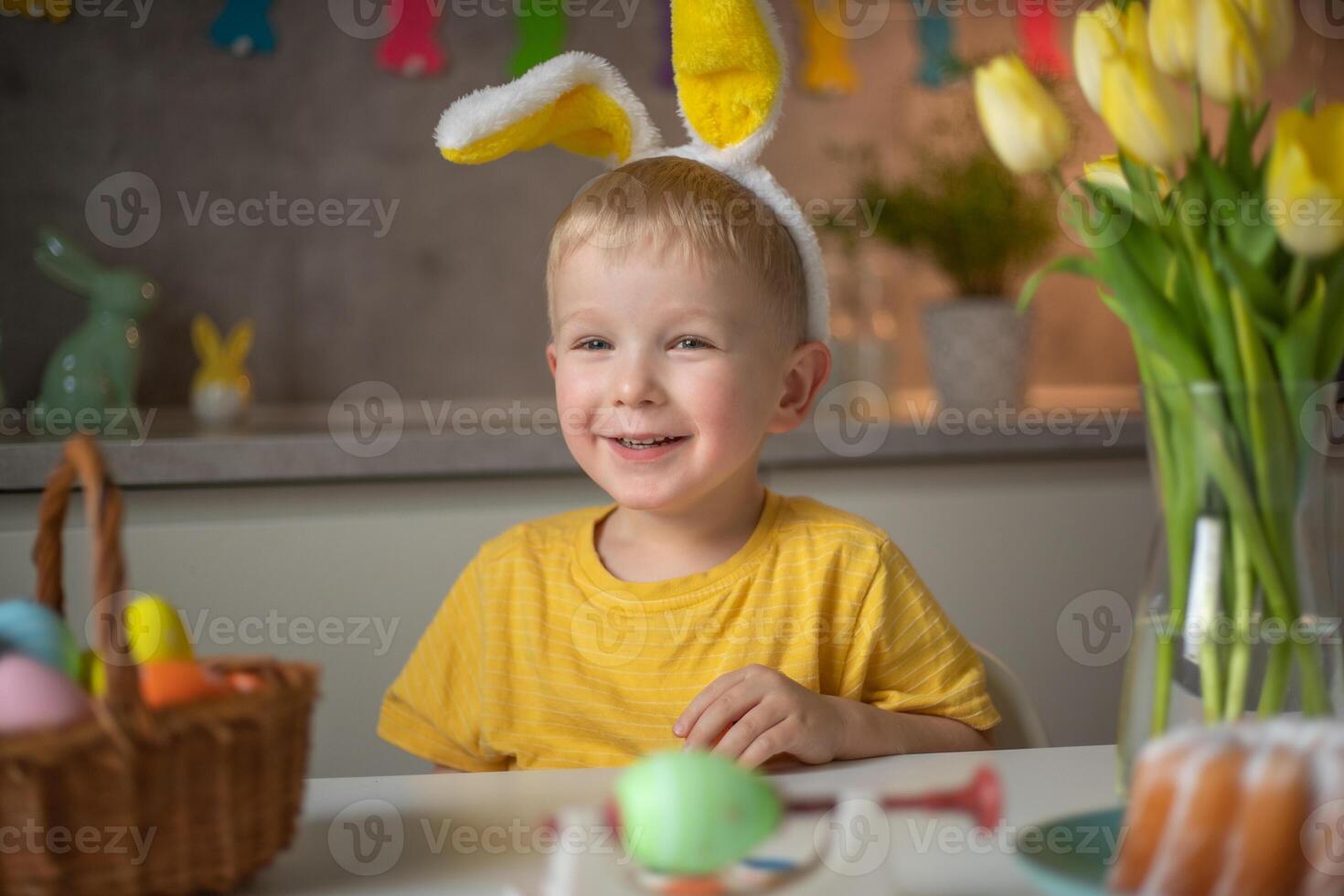 Emotional portrait of a cheerful little boy wearing bunny ears on Easter day who laughs merrily, plays with colorful Easter eggs sitting at a table in the kitchen photo