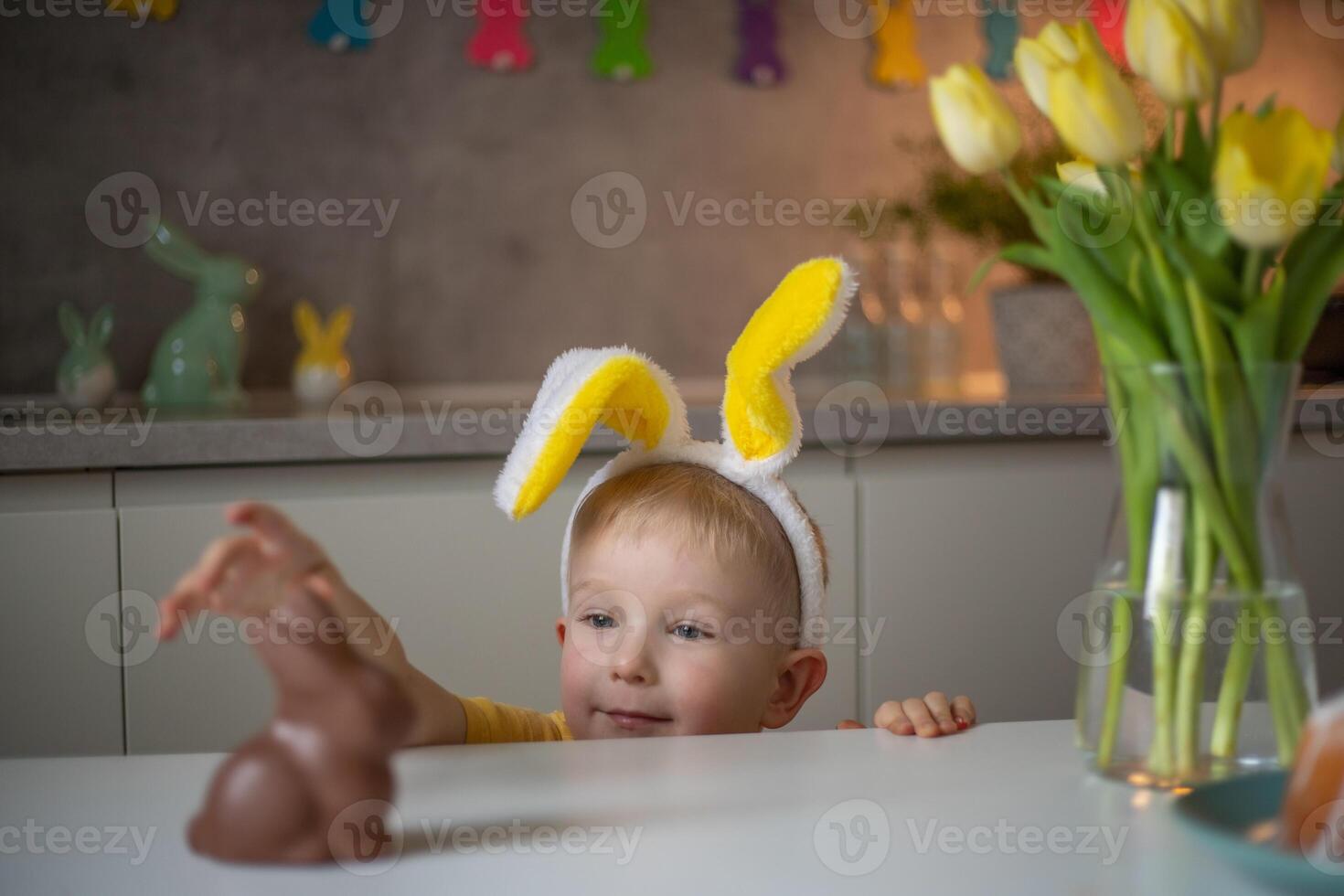 A cute little boy wearing bunny ears on Easter day wants to eat a chocolate easter bunny. A child plays egg hunt for Easter. Charming child celebrates Easter at home photo