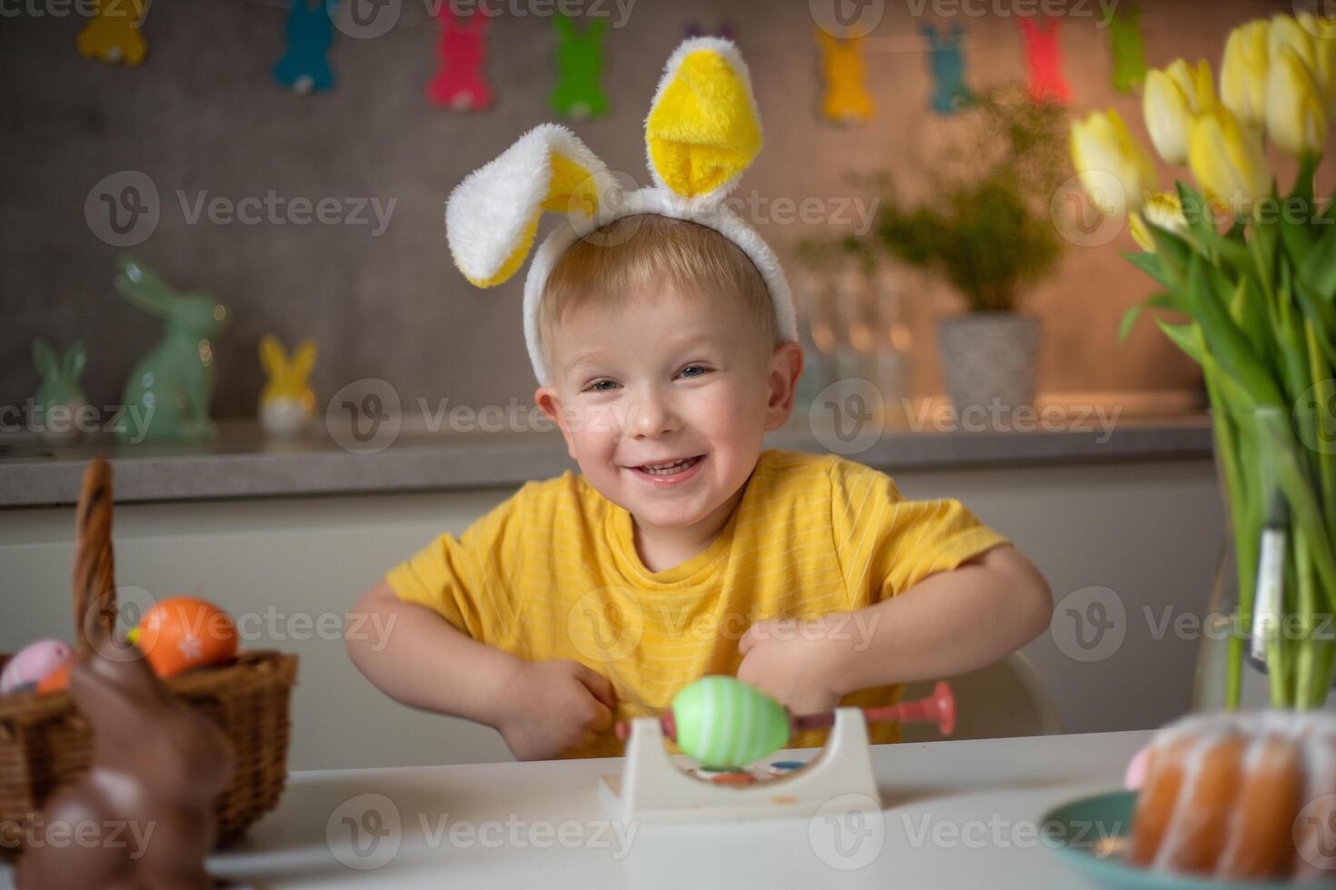 Emotional portrait of a cheerful little boy wearing bunny ears on Easter day who laughs merrily, plays with colorful Easter eggs sitting at a table in the kitchen photo