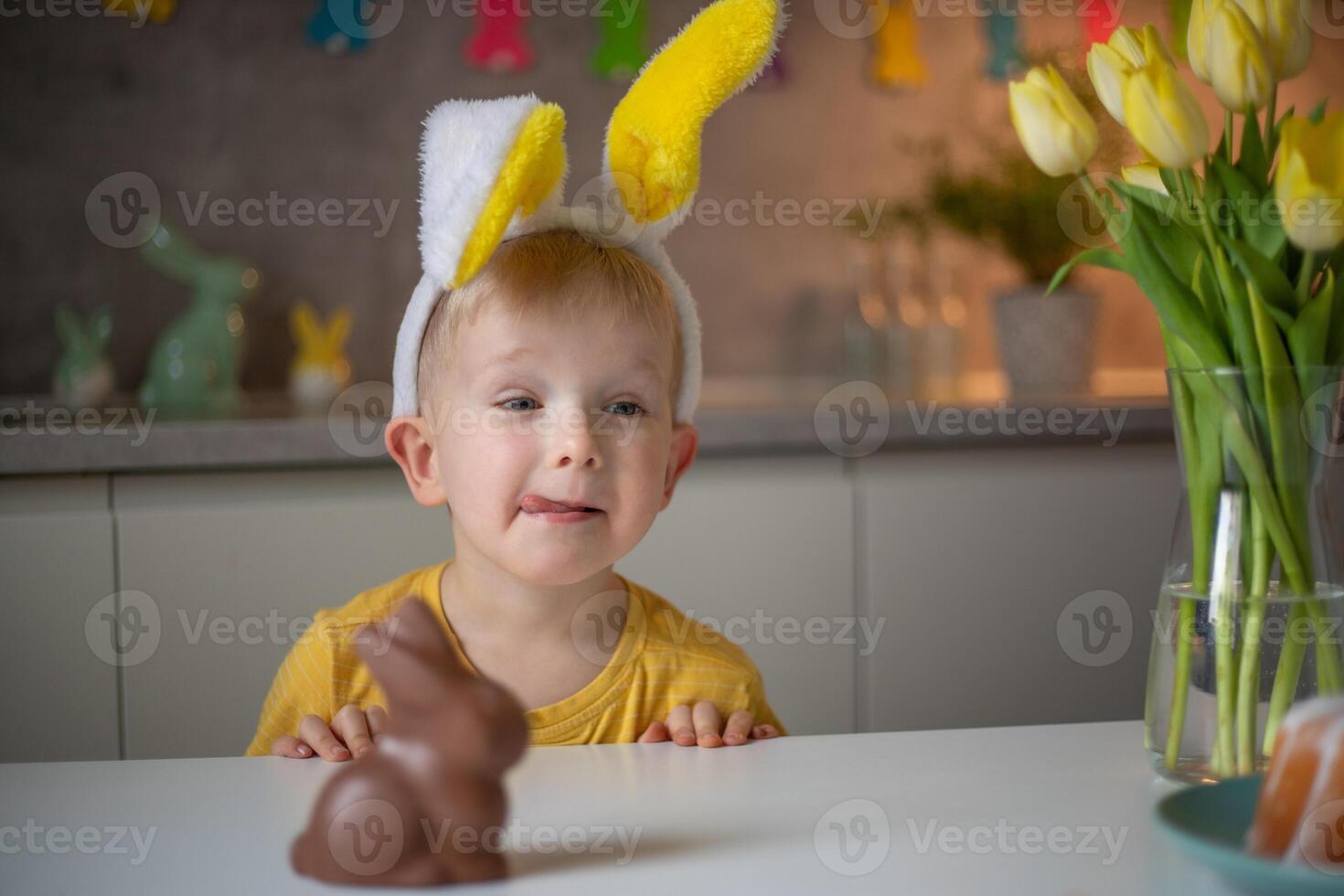 A cute little boy wearing bunny ears on Easter day wants to eat a chocolate easter bunny. A child plays egg hunt for Easter. Charming child celebrates Easter at home photo