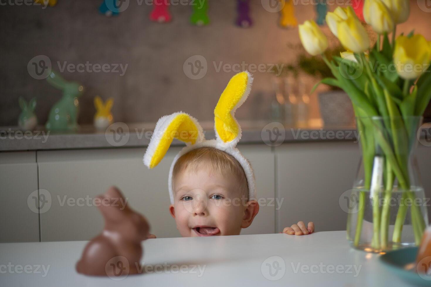 A cute little boy wearing bunny ears on Easter day wants to eat a chocolate easter bunny. A child plays egg hunt for Easter. Charming child celebrates Easter at home photo