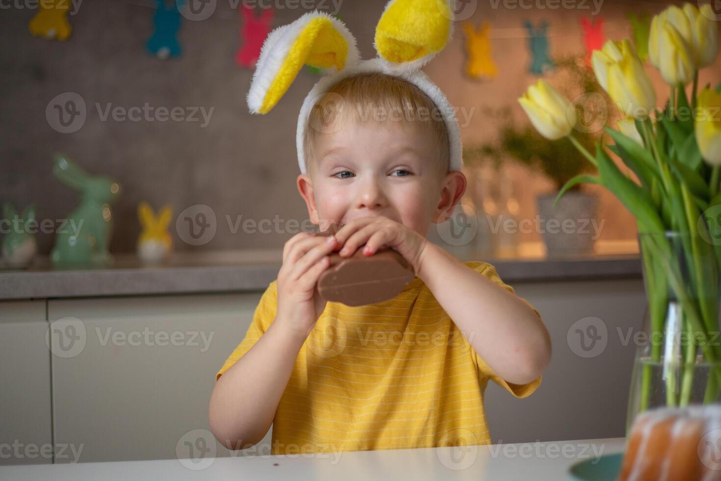 A cute little boy wearing bunny ears on Easter day is eating a chocolate Easter bunny. A child plays egg hunt for Easter. Charming child celebrates Easter at home photo