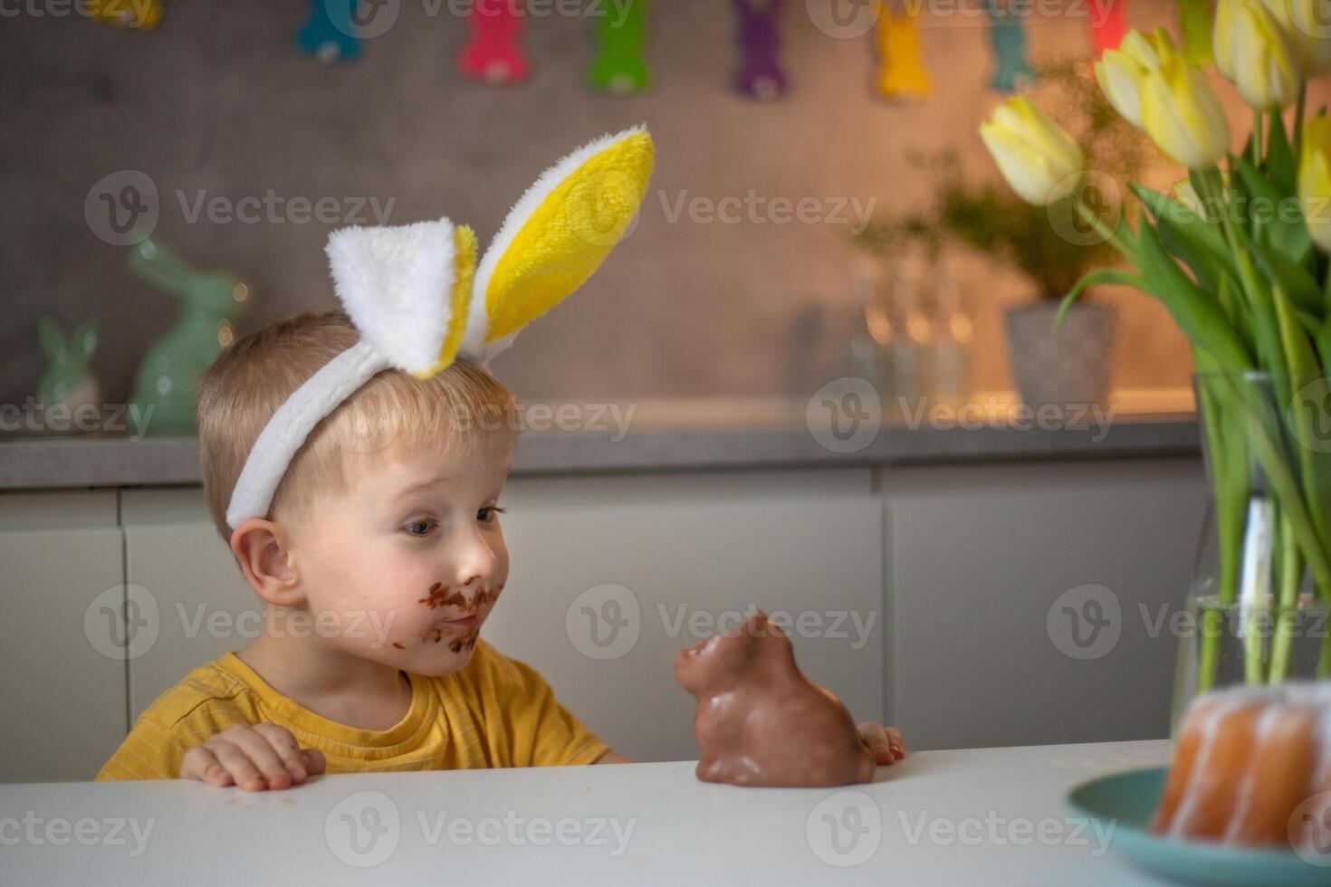 A cute little boy wearing bunny ears on Easter day is eating a chocolate Easter bunny. A child plays egg hunt for Easter. Charming child celebrates Easter at home photo