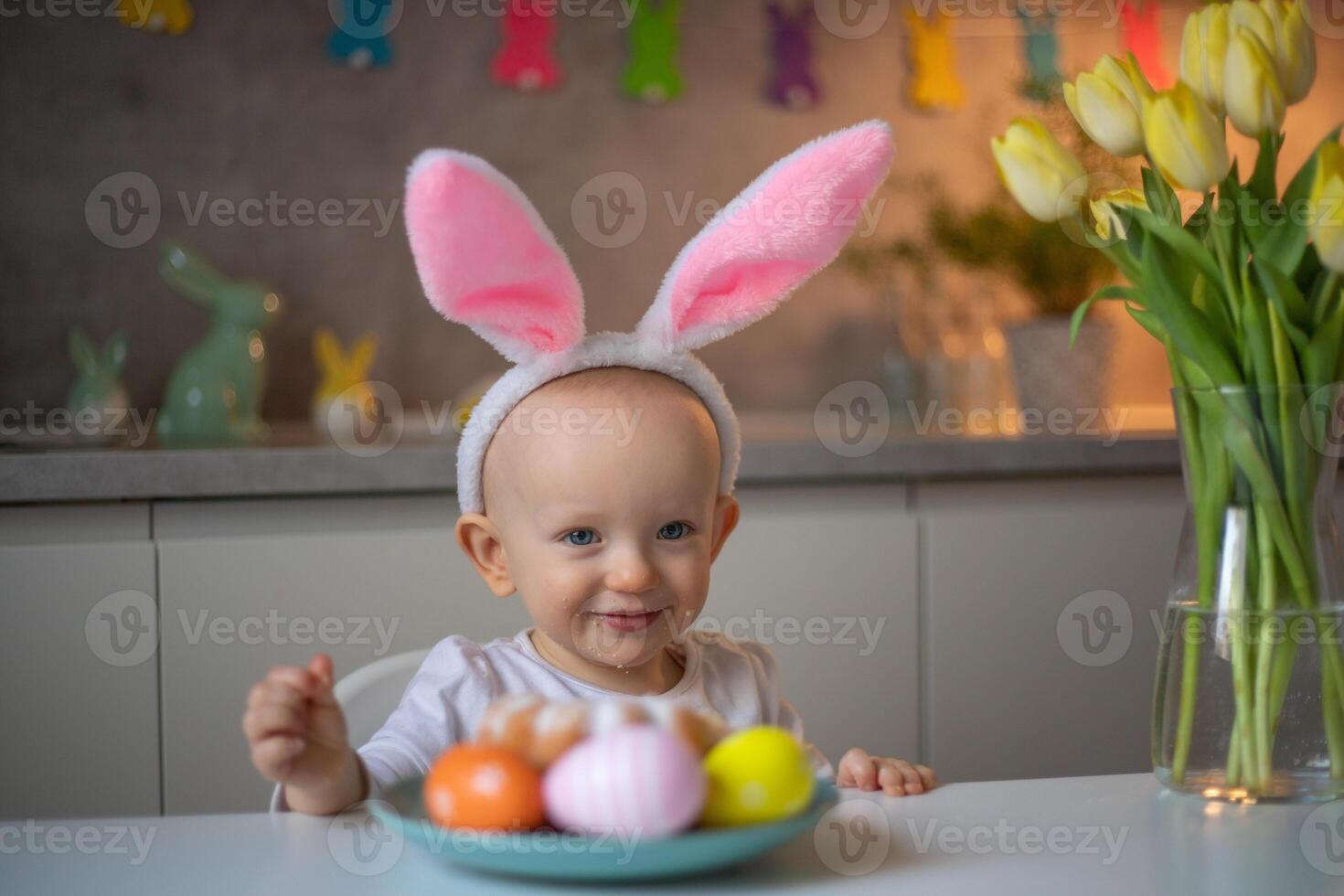 happy cute little baby girl wearing bunny ears on easter day sitting at the table in the kitchen photo