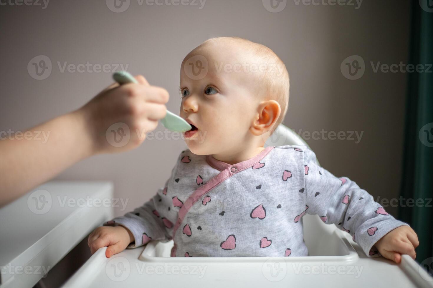 Baby girl eating blend mashed food sitting, on high chair, mother feeding child, hand with spoon for vegetable lunch, baby weaning, first solid food for young kid photo