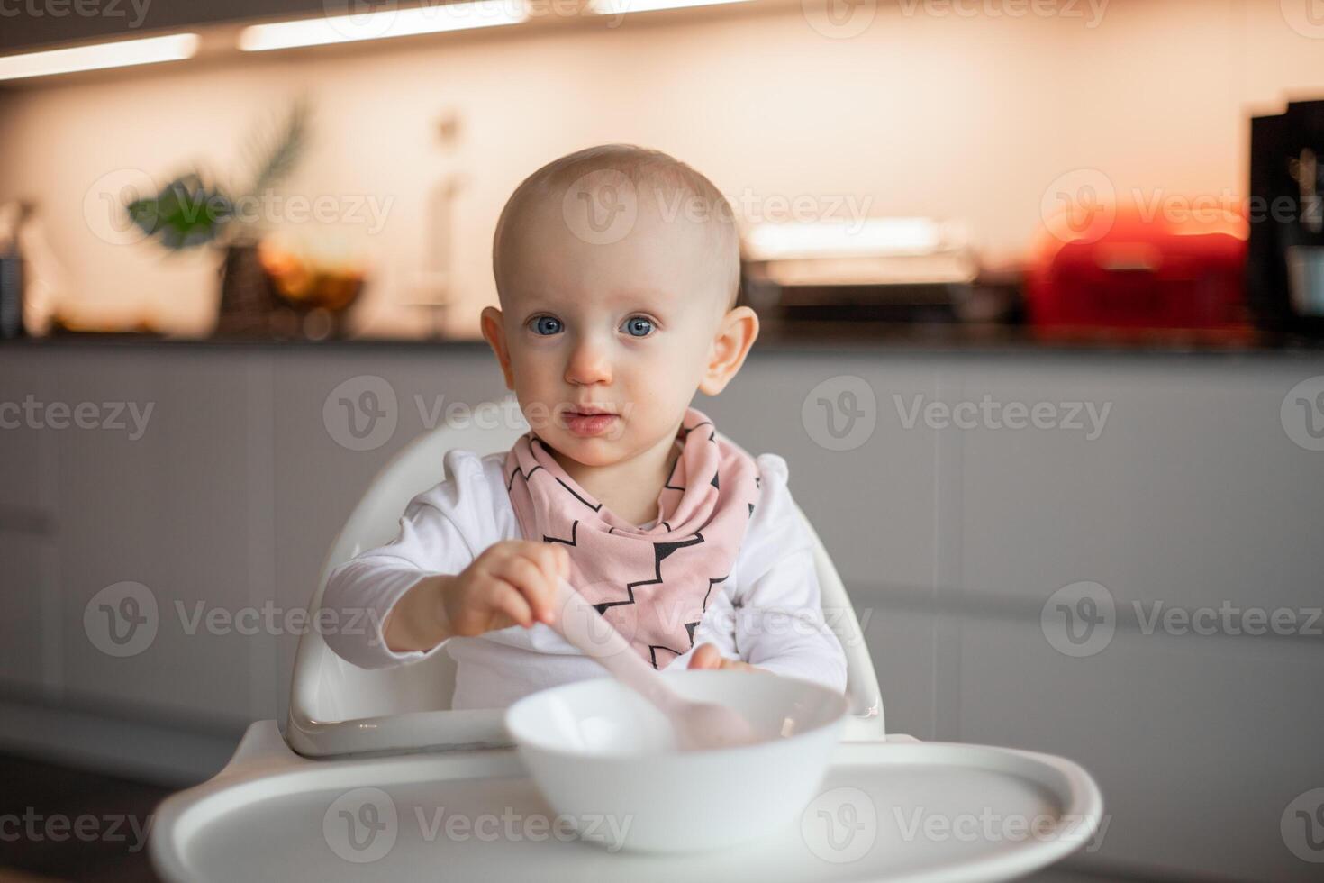 Happy little baby eats on his own with a spoon while sitting in a high chair photo