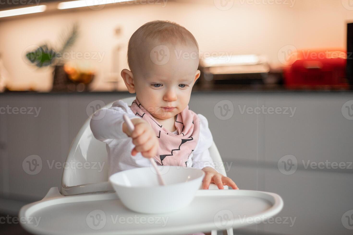 Happy little baby eats on his own with a spoon while sitting in a high chair photo