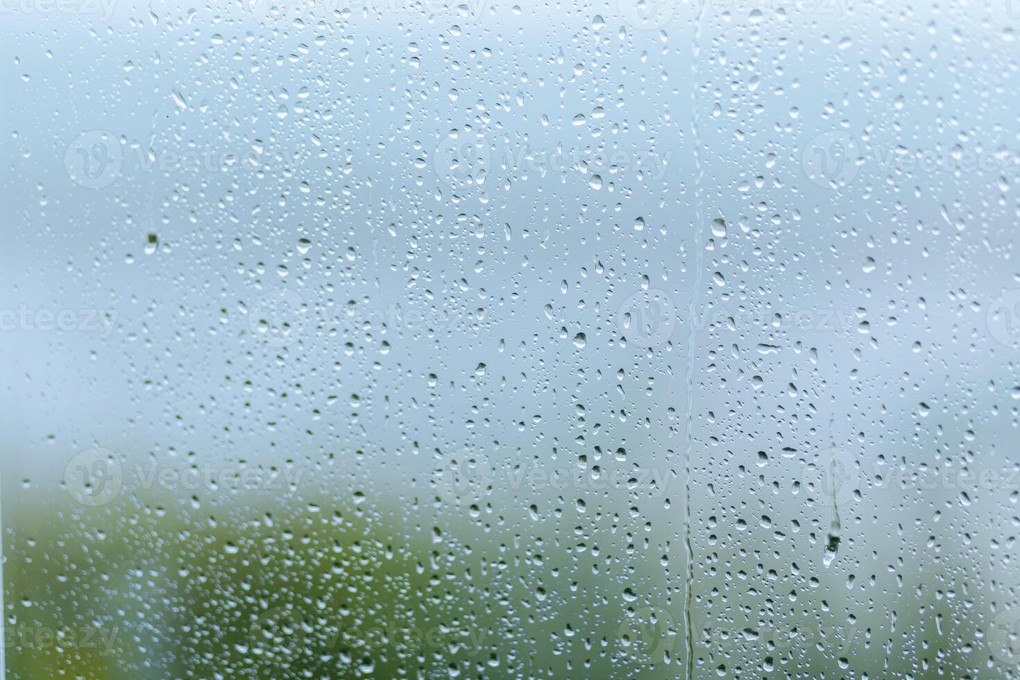 Rainy day with droplets on a windowpane, creating a blurred view of greenery outside during overcast weather photo